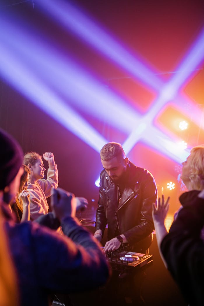 DJ in black leather jacket mixing music at a lively club party with vibrant lights.
